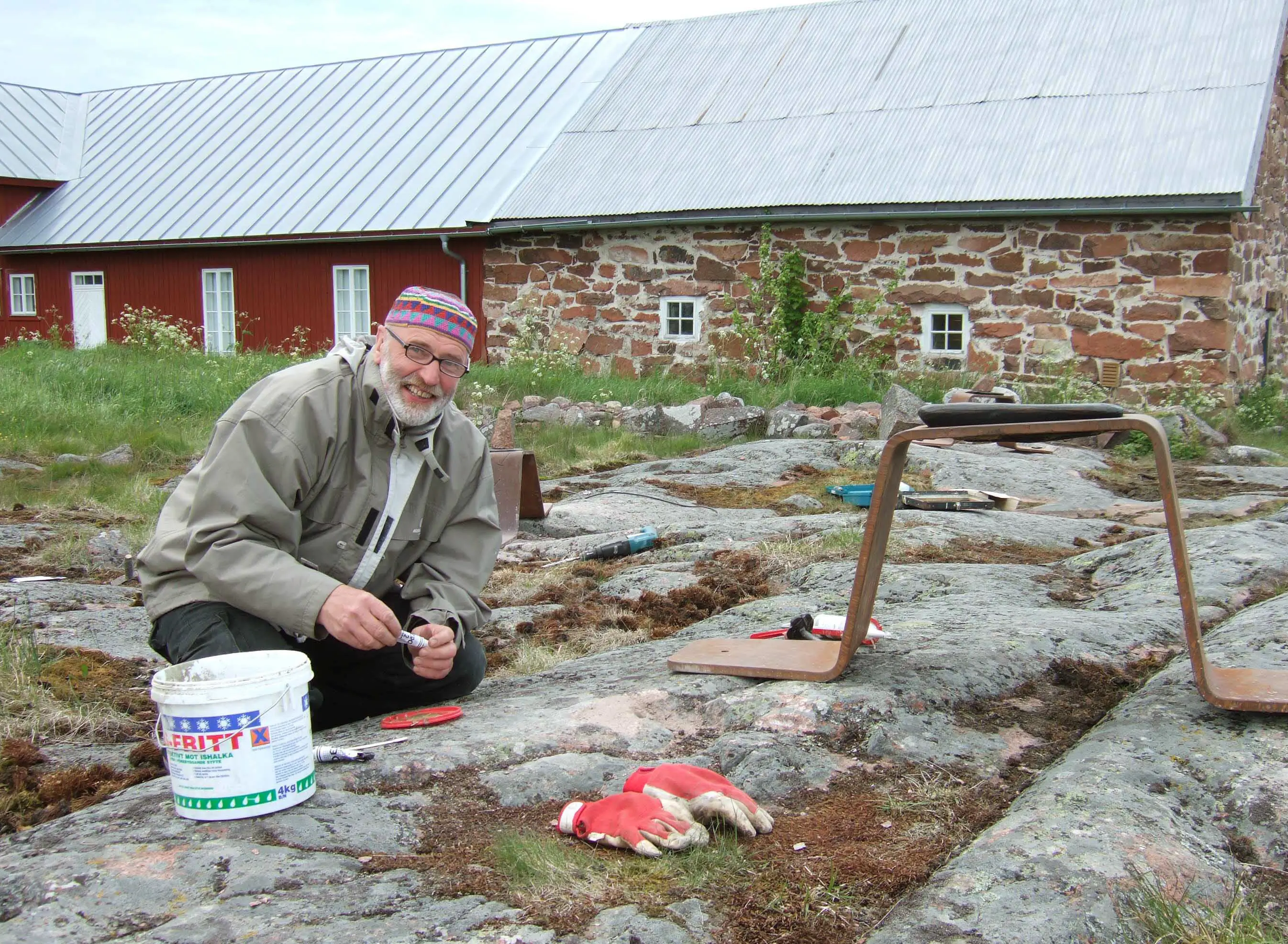 Foto som visar skulptören Jüri Ojaver sittandes med utrustning samt järndelar på mossig, stenig mark. Jüri har en tunn mössa, vitt kortklippt skägg och ljusgrå vindtygsjacka. I bakgrunden syns en långsträckt rödmålad träbyggnad