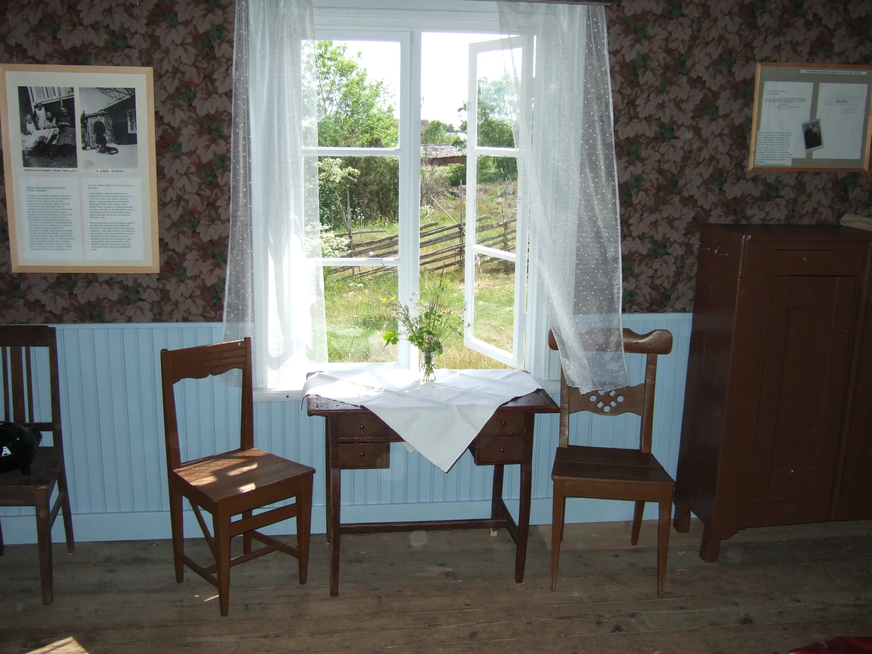 Fotografi av Sjöbloms emigrant- och torparmuseum, interiör. Bilden visar ett mörkbrunt äldre bord med lådor prytt av en broderad vit duk, tillsammans med två stolar framför ett halvöppet fönster. Tunna, vita gradiner fladdrar i vinddraget. Utanför fönstret syns en vildvuxen gräsmatta och några meter bort en gärdsgård och lövträd.